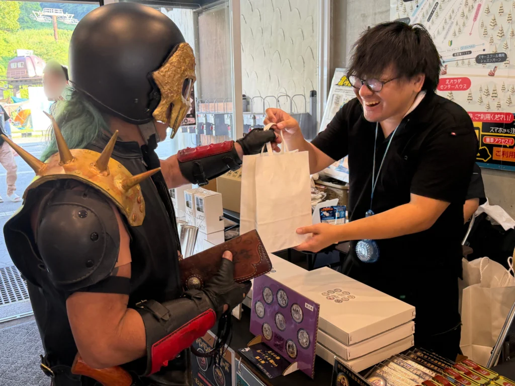 北斗の拳「ジャギ」のコスプレをしたお客様に、タク技研スタッフが笑顔で商品をお渡しする様子